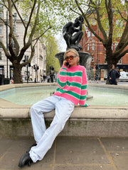 A person sitting on a fountain edge wearing a pink and green striped mohair jumper with white trousers.