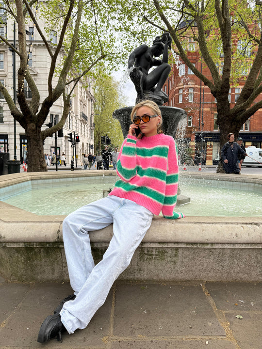 A person sitting on a fountain edge wearing a pink and green striped mohair jumper with white trousers.